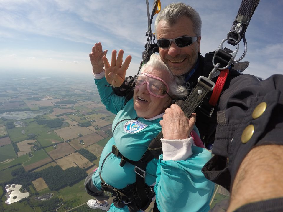 Bo Babovic smiles at the camera 4000 feet in the air suspended under canopy with his tandem student, Anna Mae