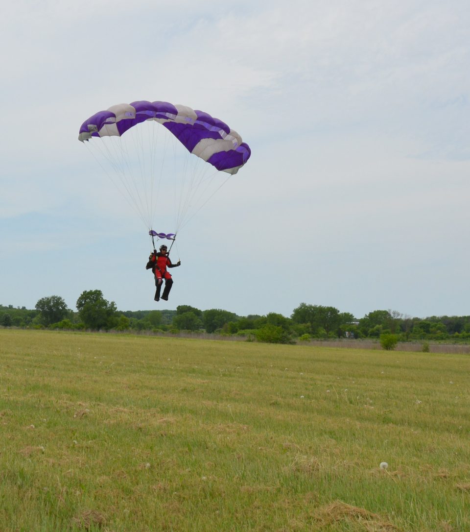 A skydiver with an open parachute about to land in a large grassy area. 
