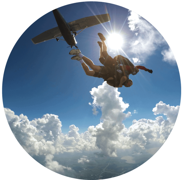 The sun shines bright, puffy white clouds, and an airplane are the background to a tandem skydiving pair follow exit.