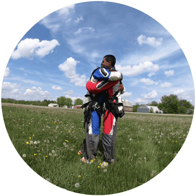 Tandem students hug in a field filled with lush, green grass and dandelions following their skydive.