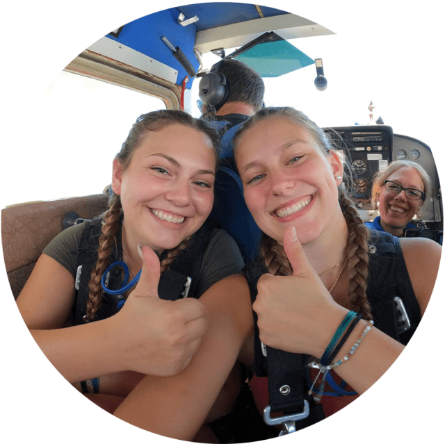Two female tandem skydivers give a thumbs up in their airplane on the way to altitude.