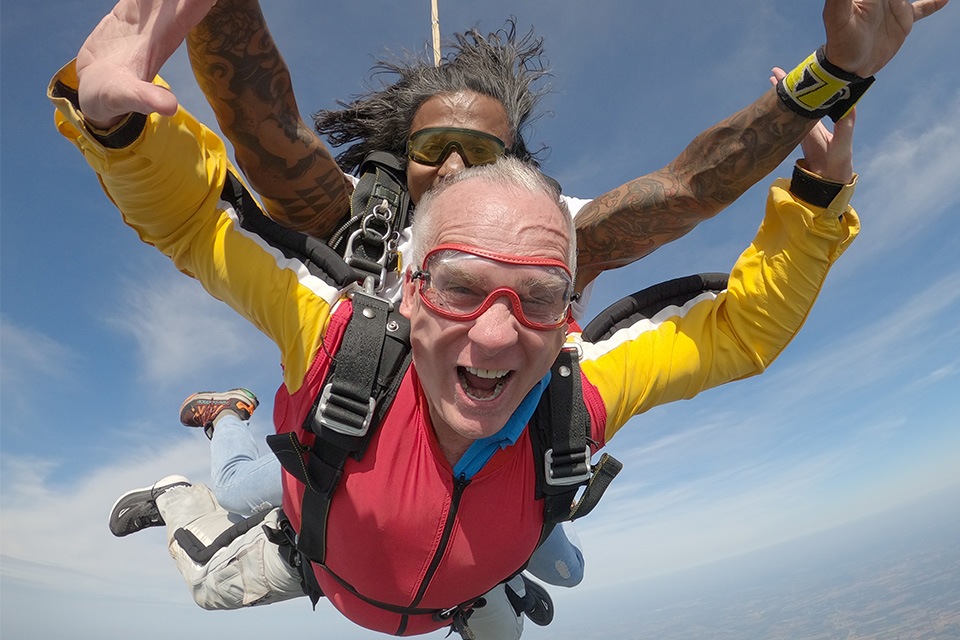 Older man with red goggled and a red and yellow jumpsuit during a tandem freefall skydive with his arms outstretched